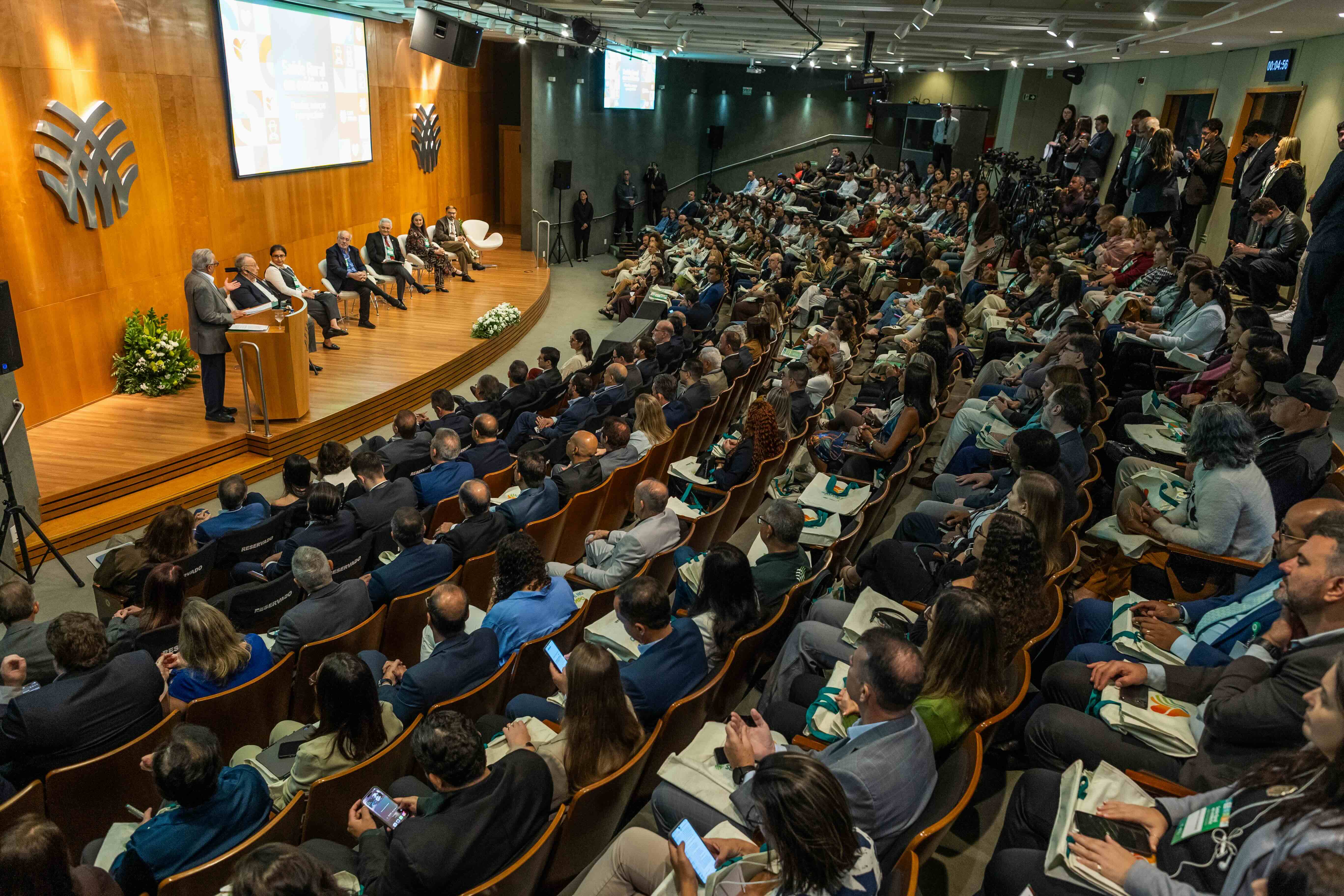 Vista ampla de um auditório lotado durante o encontro, com palestrantes sentados no palco e um apresentador no púlpito.